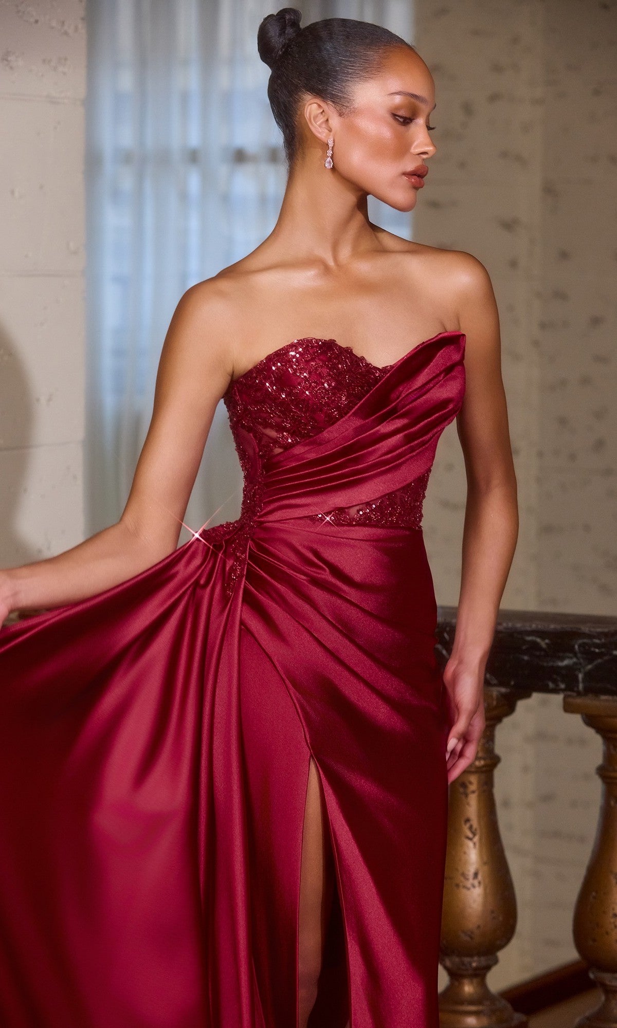 A woman wearing the Ladivine J887 Side-Drape Strapless Long Prom Gown in burgundy satin with a sequined bodice and high slit stands by a staircase, holding part of her elegant evening gown.